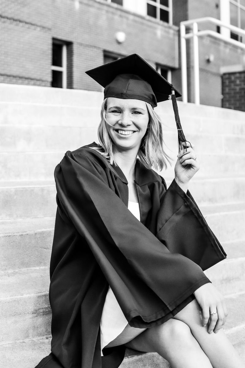 Monochrome image of a female in a cap and gown and sitting on the stairs for senior portraits.