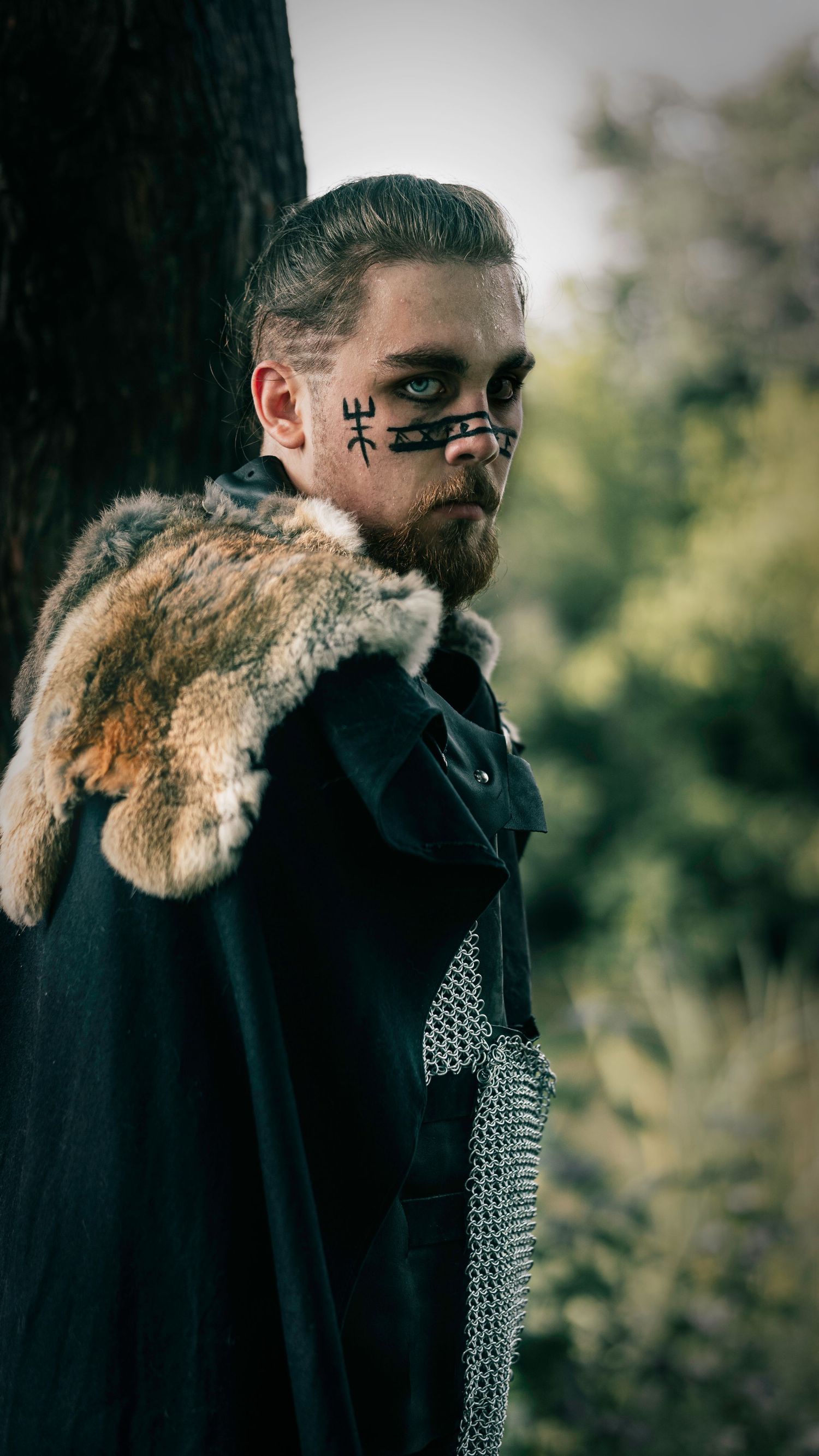 Medieval warrior wearing fur-trimmed cloak and chainmail with face paint stands against blurred nature background.