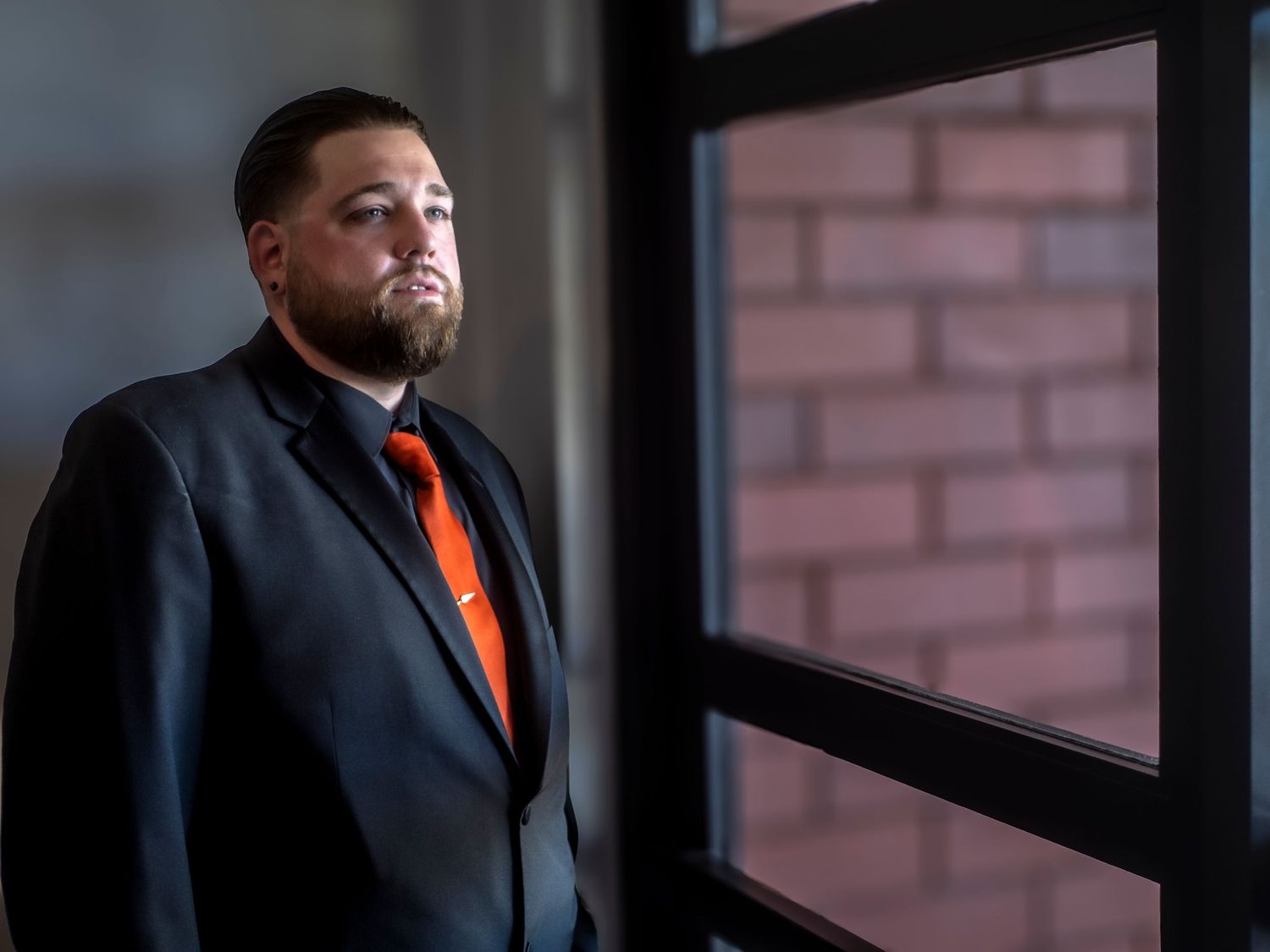 Professional wearing a dark suit with orange tie gazes thoughtfully through window in corporate setting.
