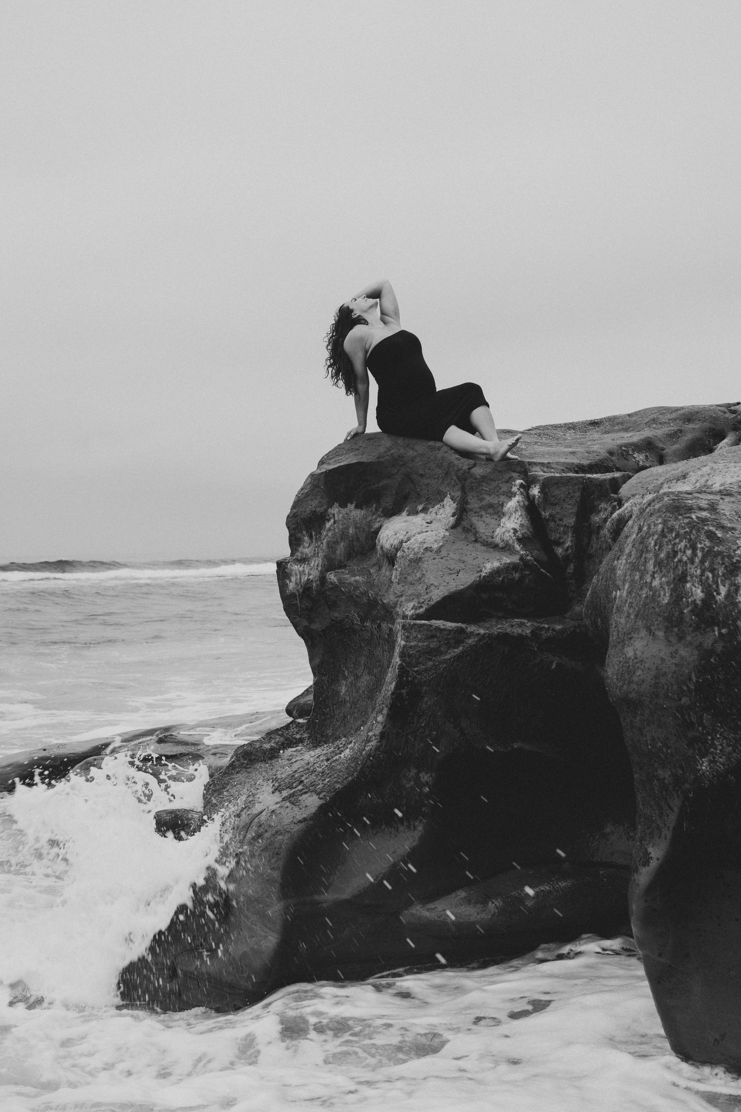 A silhouette rests dramatically on coastal rocks as waves crash against the cliffs in a moody black and white scene.