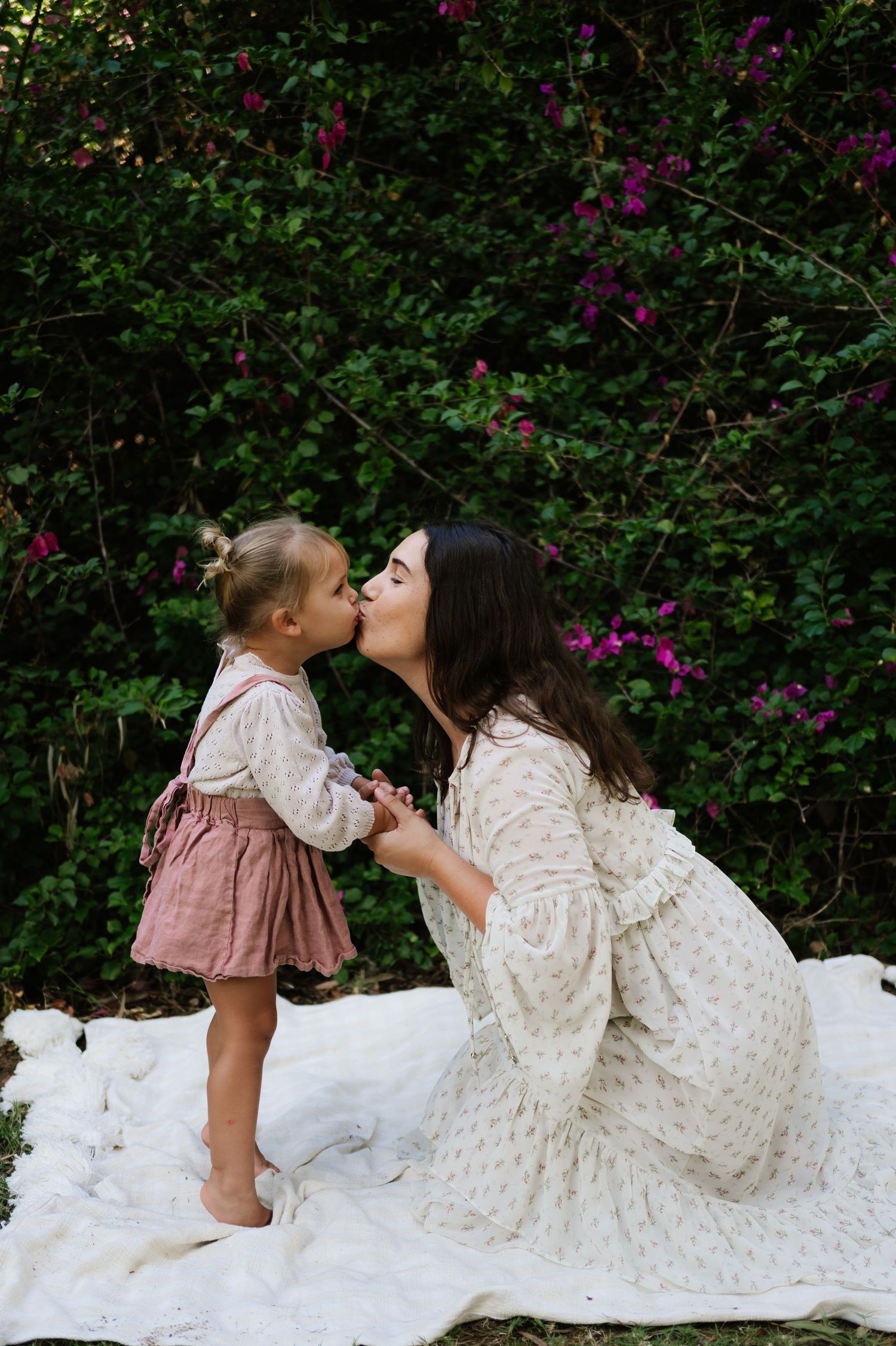 A tender moment shared between two people in white dresses against a backdrop of purple flowering bushes.