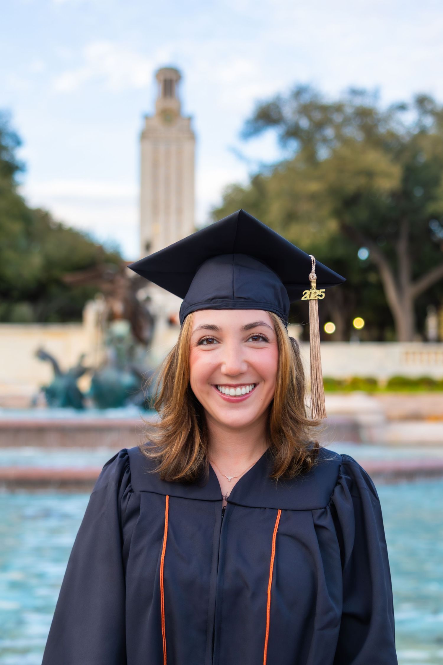 Graduate in cap and gown poses in front of the UT Austin Tower and Littlefield Fountain during graduation photos.
