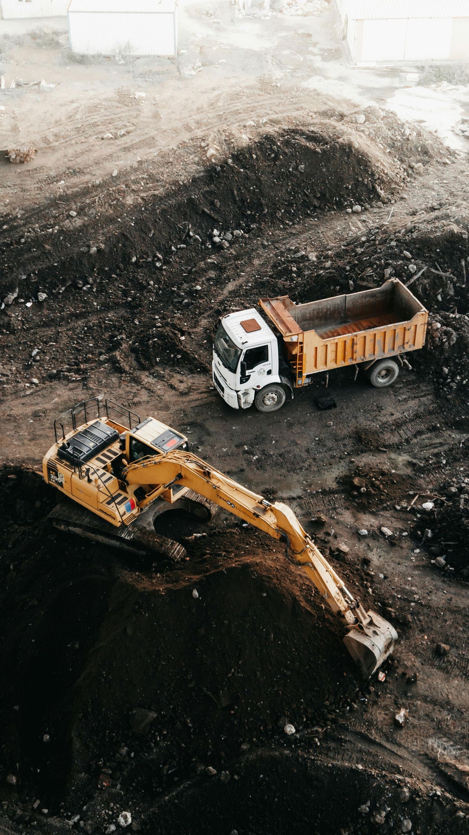 Yellow excavator and white dump truck working on dark soil at a construction site.