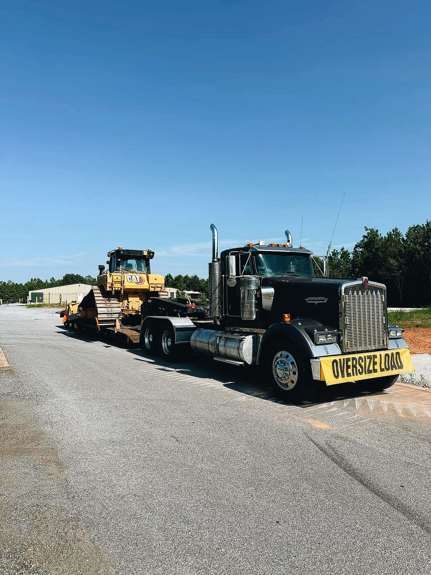 Black Kenworth semi truck hauling heavy equipment with Oversize Load banner on a sunny day.