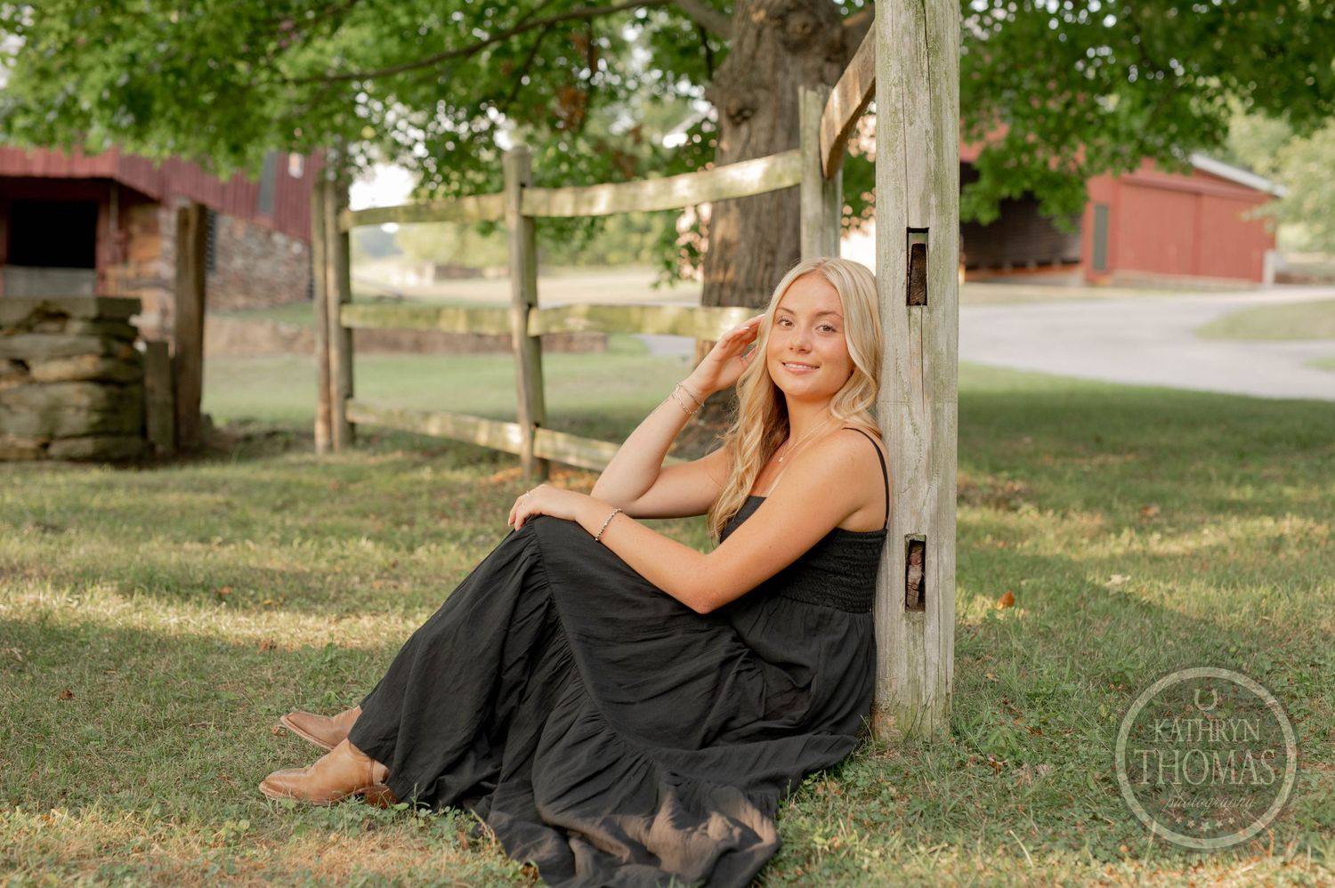 A woman in a black dress relaxes against a wooden post in a serene country setting.