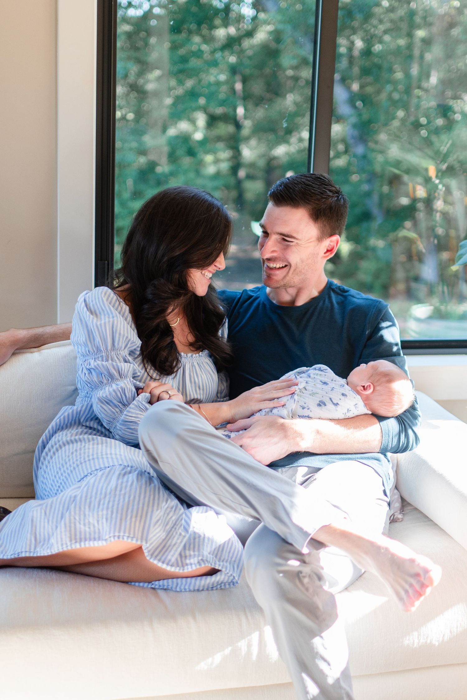 A couple lounges on a white couch in bright natural light while holding their newborn baby.