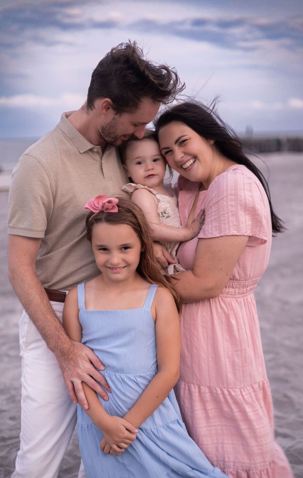 A family wearing pastel pink and blue outfits embraces lovingly on a cloudy beach.