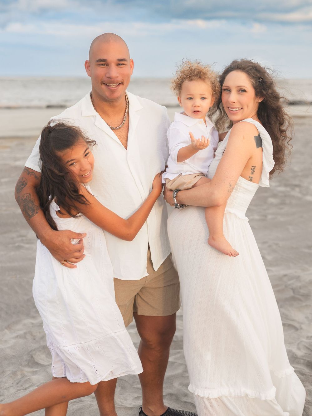 A family wearing white outfits poses together for casual beach portraits at sunset along the shoreline.