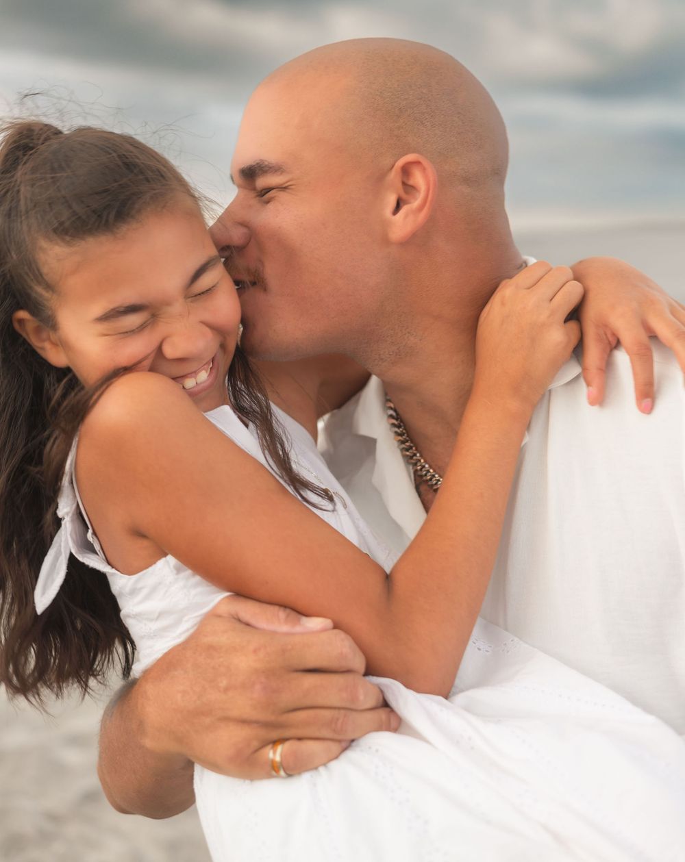 A loving couple embraces and shares a tender kiss on a beach with cloudy skies in the background.