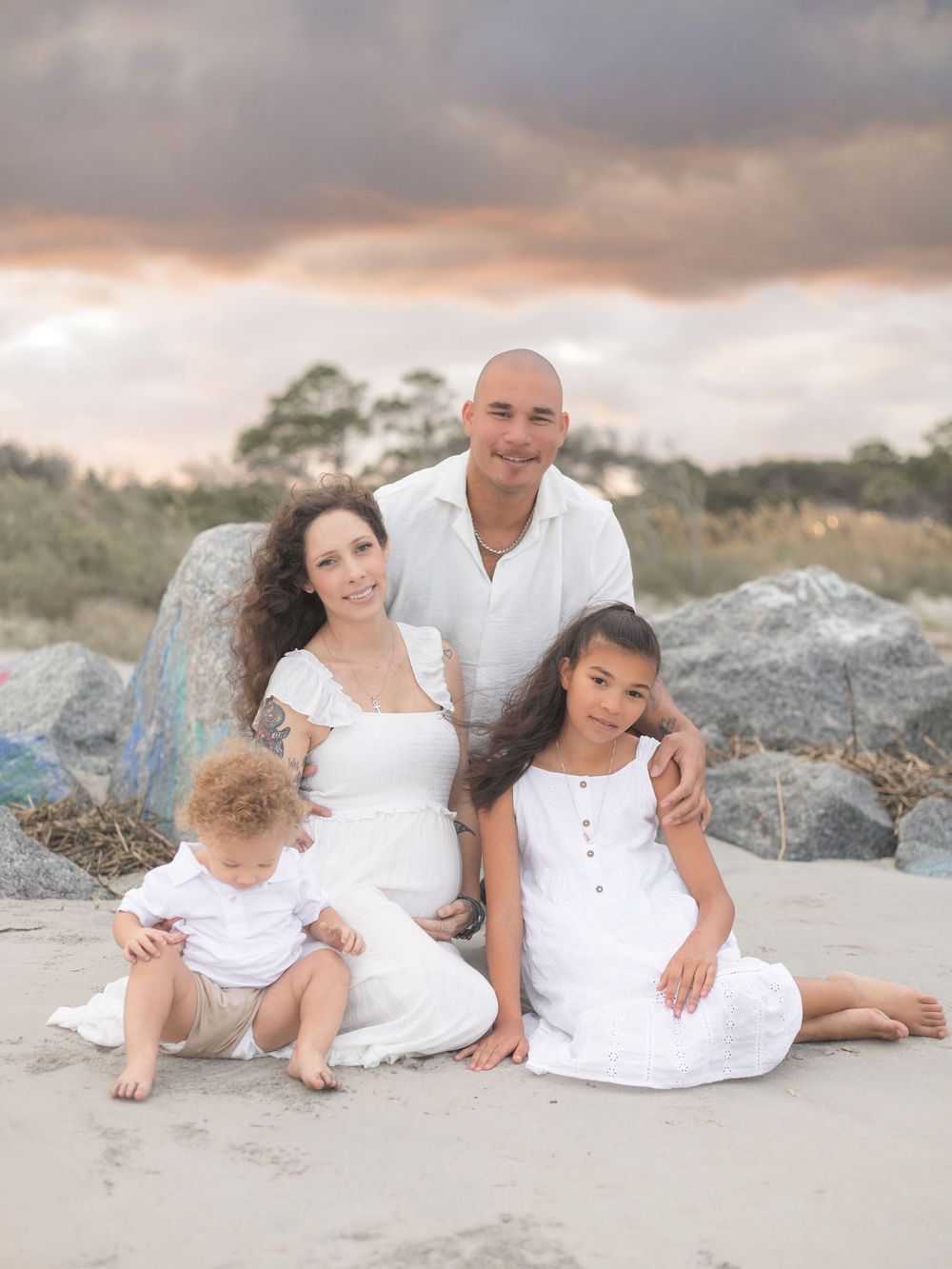 Family wearing white outfits poses together on sandy beach at sunset with rocky coastline in background.