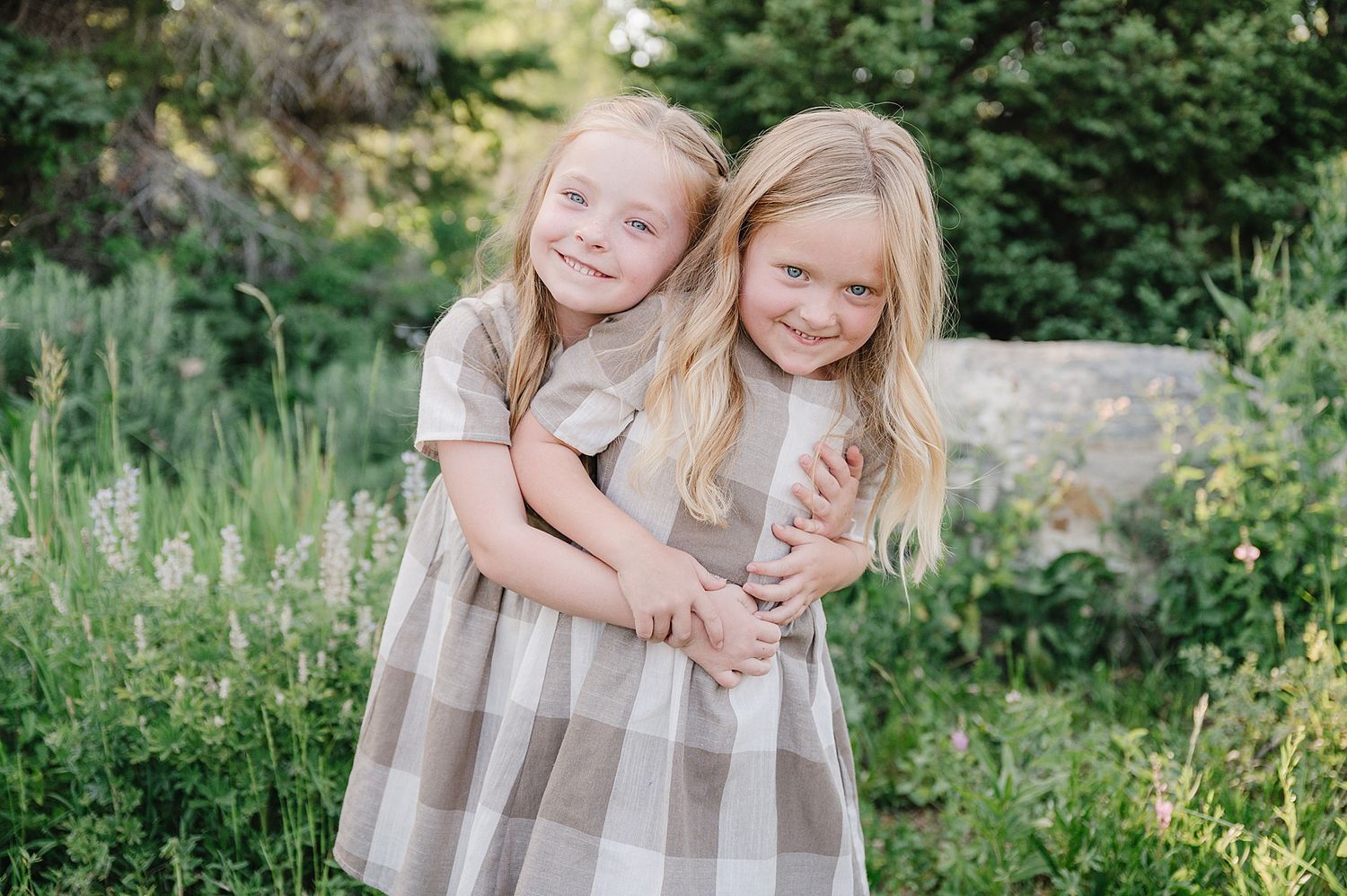 Two children in matching plaid dresses embrace while smiling outdoors among greenery and rocks.