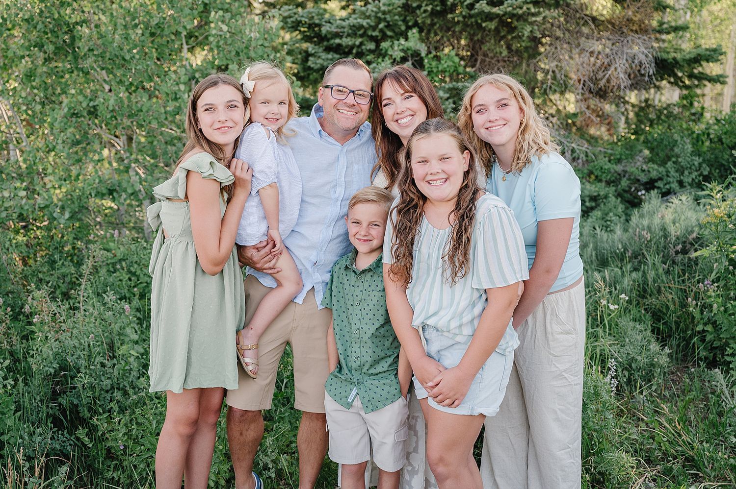 Family members gather for a portrait outdoors against a backdrop of green foliage wearing summer casual clothing.
