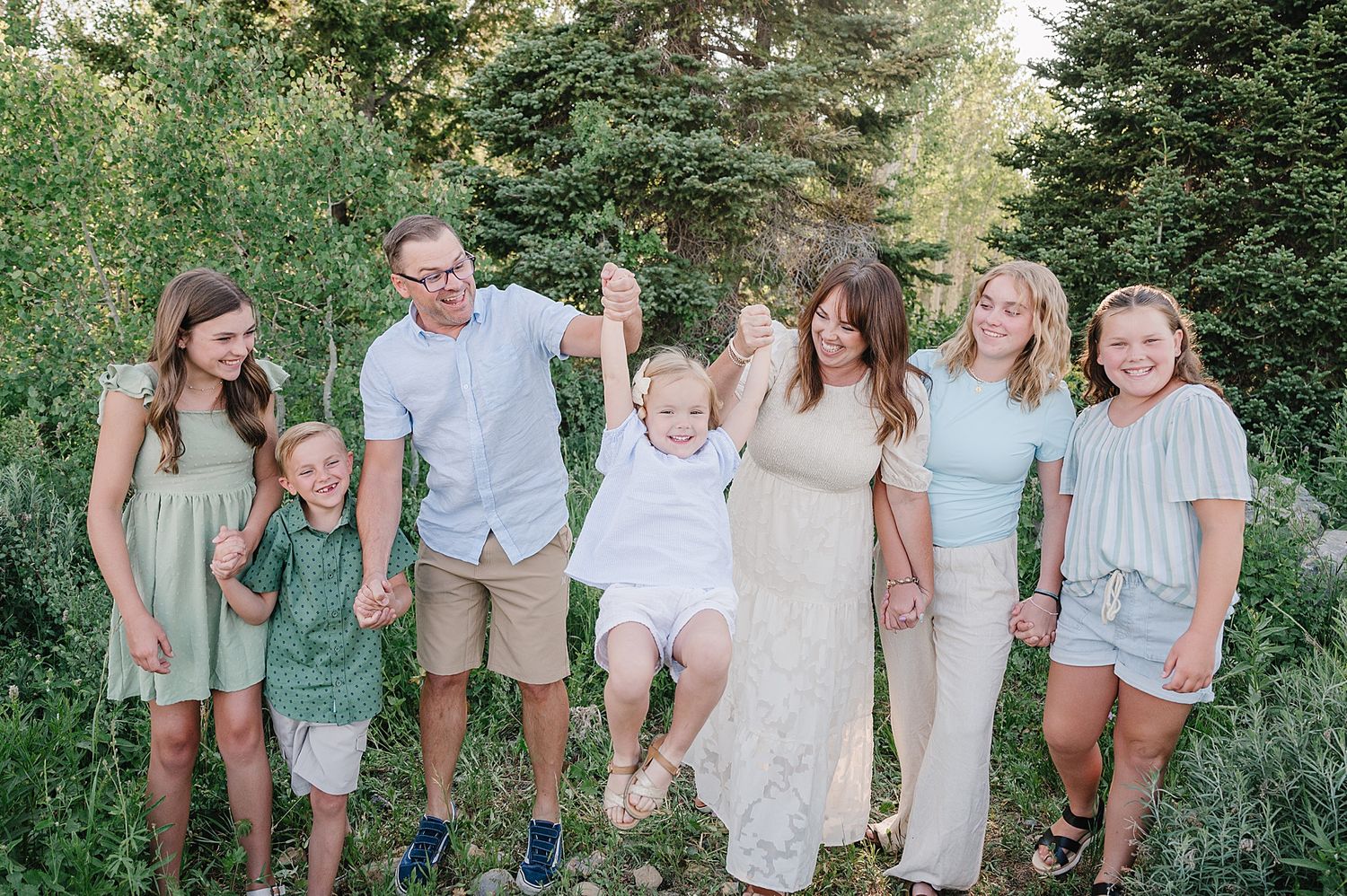 A group poses together outdoors in casual summer attire against a backdrop of evergreen trees.