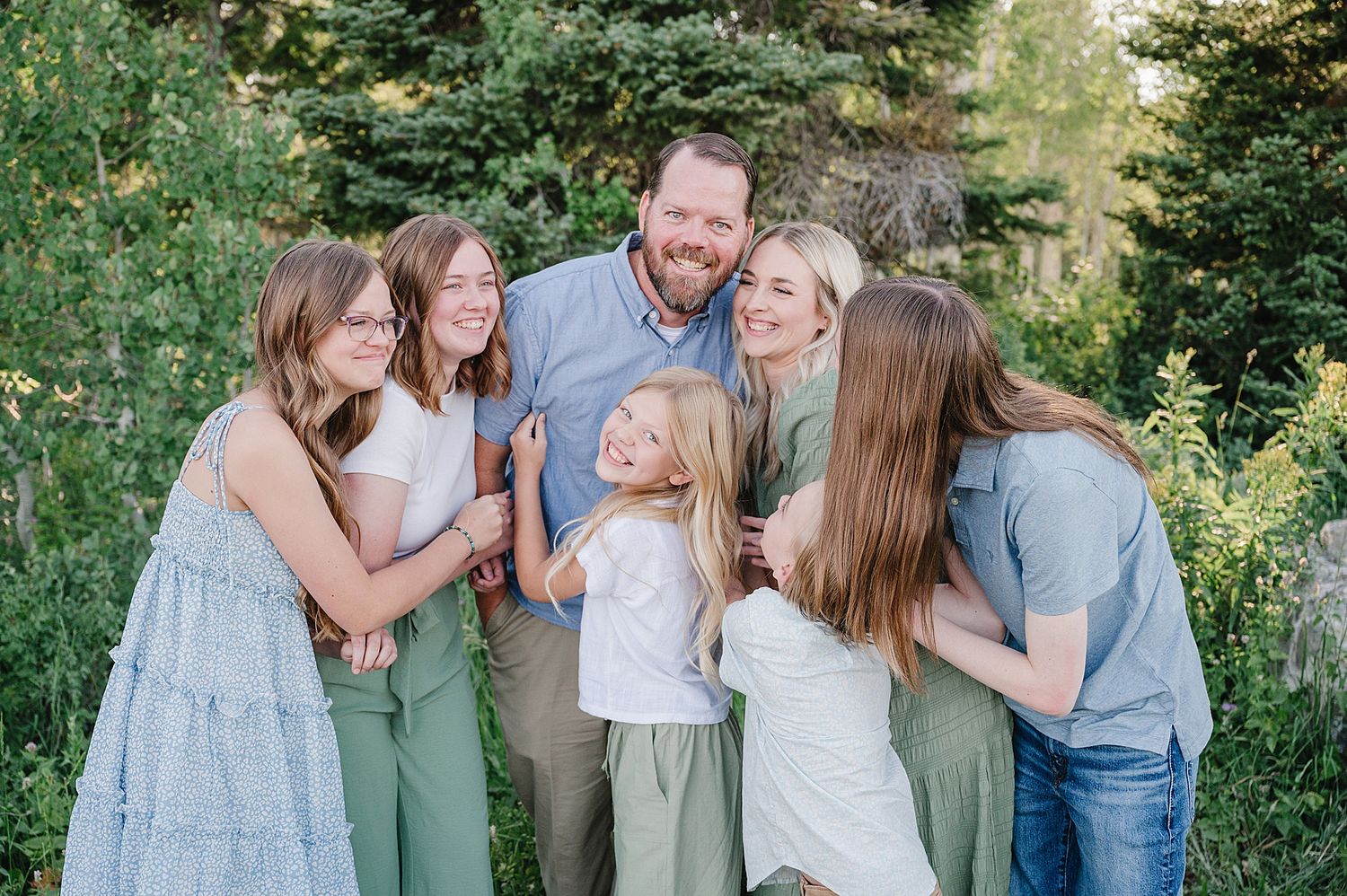 A joyful outdoor family portrait in casual clothing surrounded by green foliage on a sunny day.