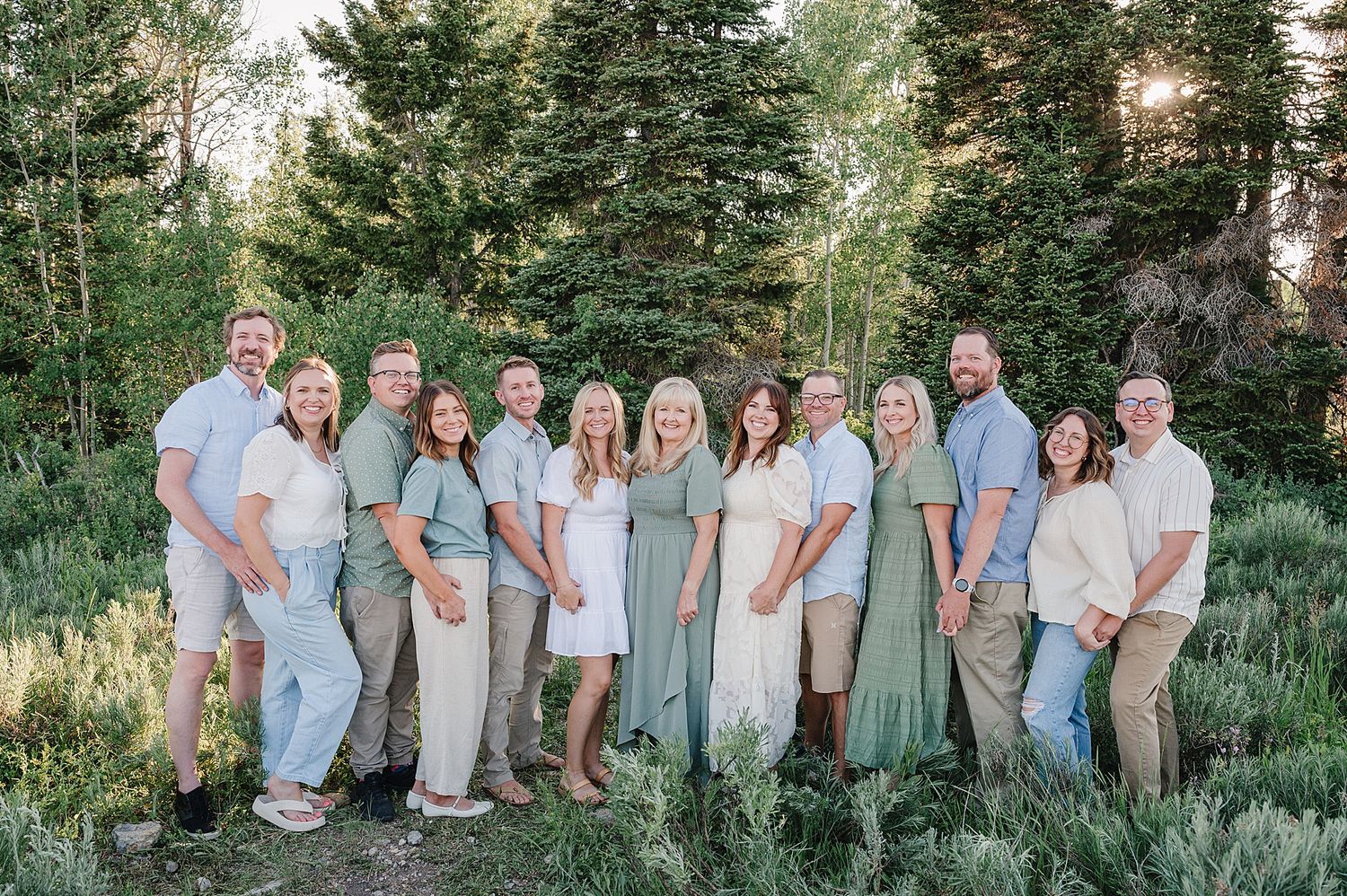 A group poses outdoors against a backdrop of evergreen trees wearing coordinated sage green and light colored outfits.