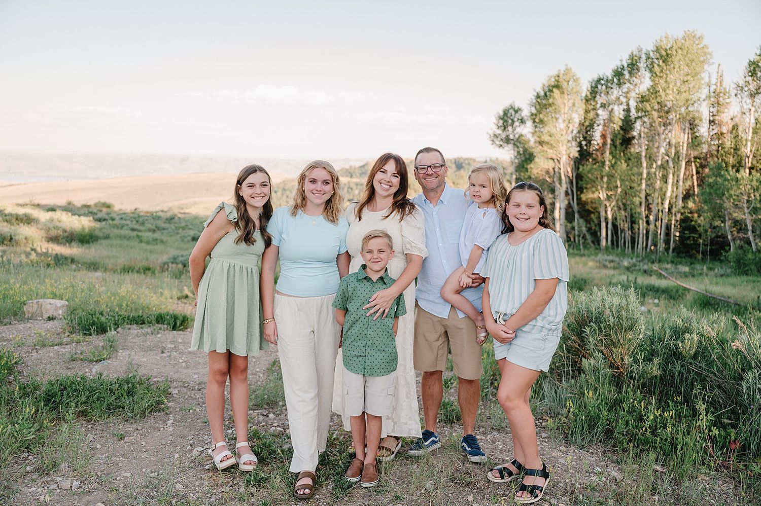 A group in coordinating pastel and neutral outfits poses together on a grassy hillside with birch trees at sunset.
