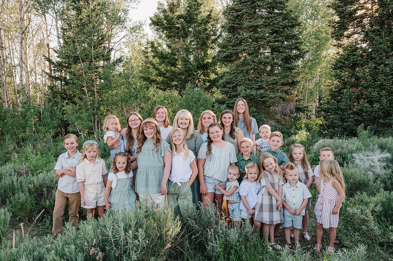 A large family gathering stands together in a wooded setting wearing matching neutral and sage colored clothing.
