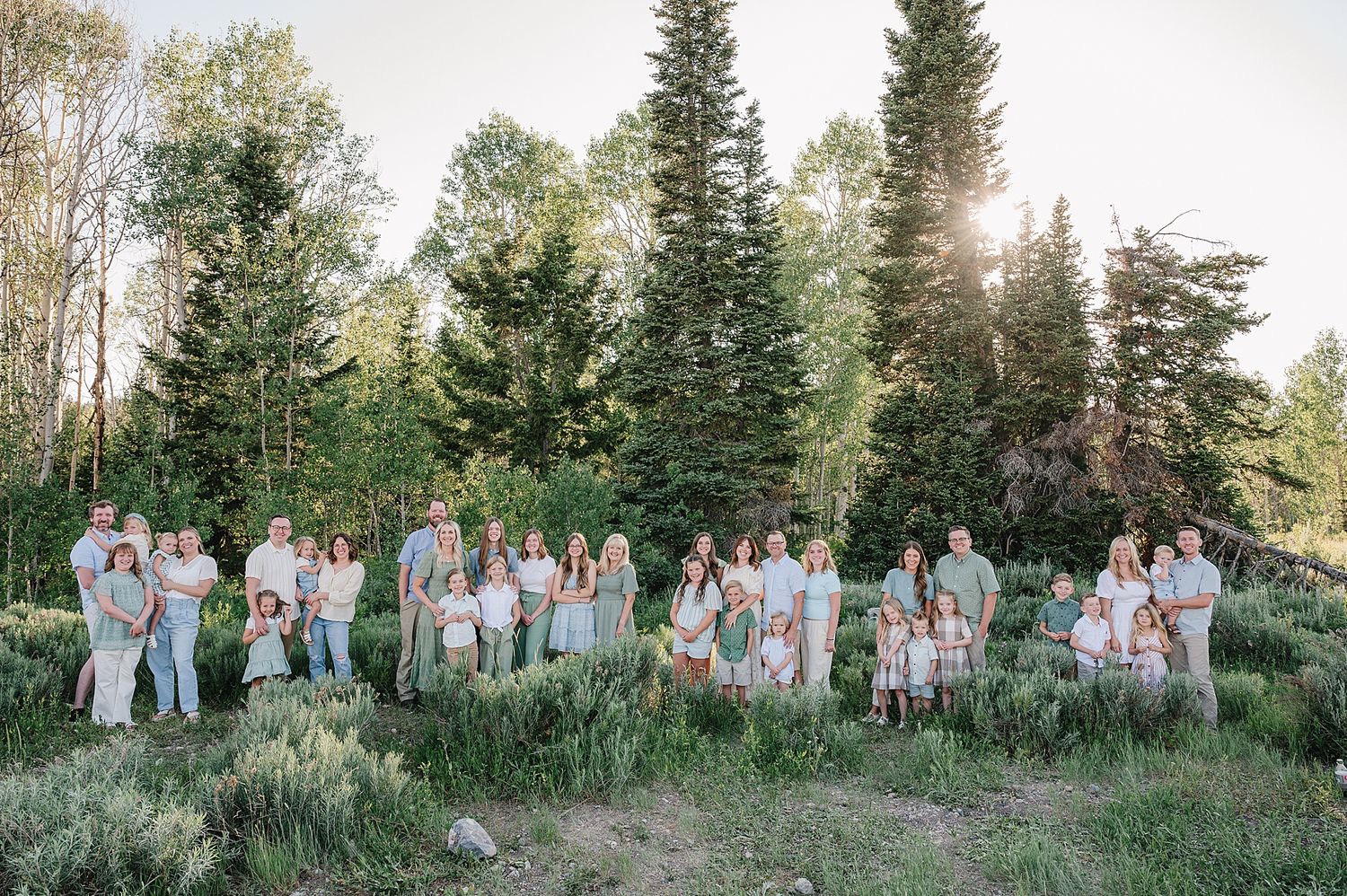 A large wedding party poses outdoors in coordinated pastel outfits against a backdrop of evergreen trees at sunset.