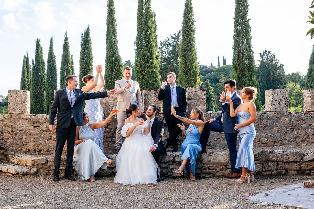 A wedding party poses casually against an ancient stone wall with cypress trees in the background at Castello di Badia in Tuscany, Italy.