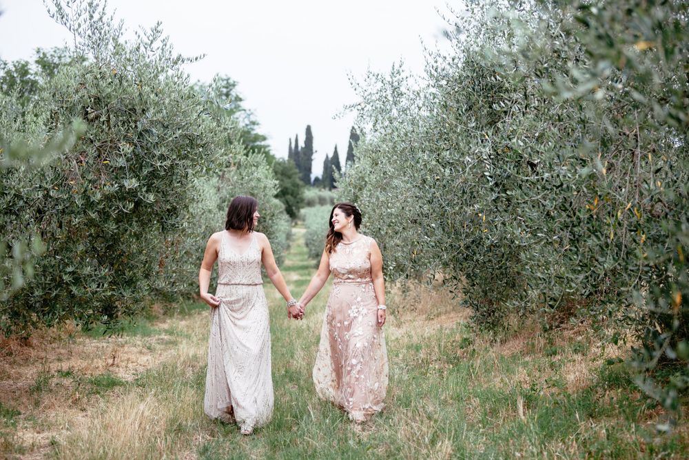 Two brides walk together through an olive grove path in soft summer light in Tuscany Italy on their wedding day.