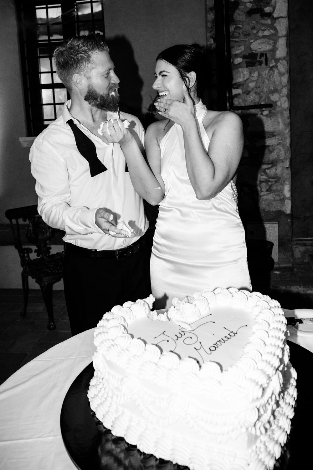 A black and white wedding photo shows a couple sharing a joyful moment while standing next to their heart-shaped wedding cake.