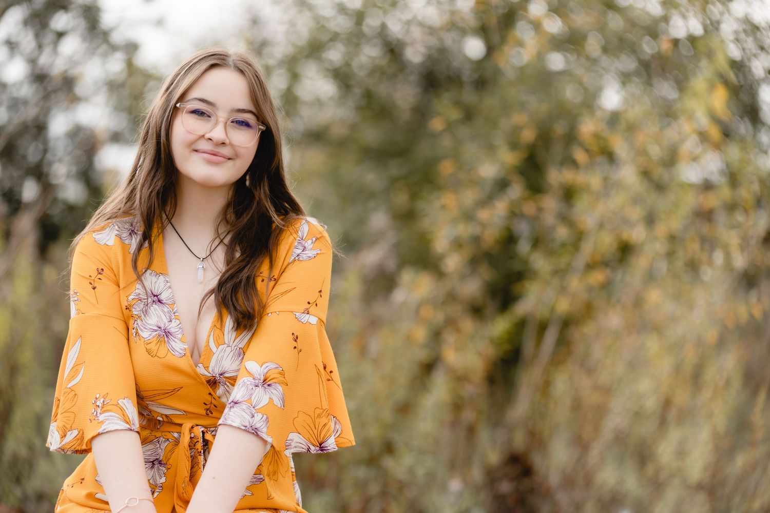 Someone in a mustard yellow floral dress smiles while posing outdoors against a blurred natural background.