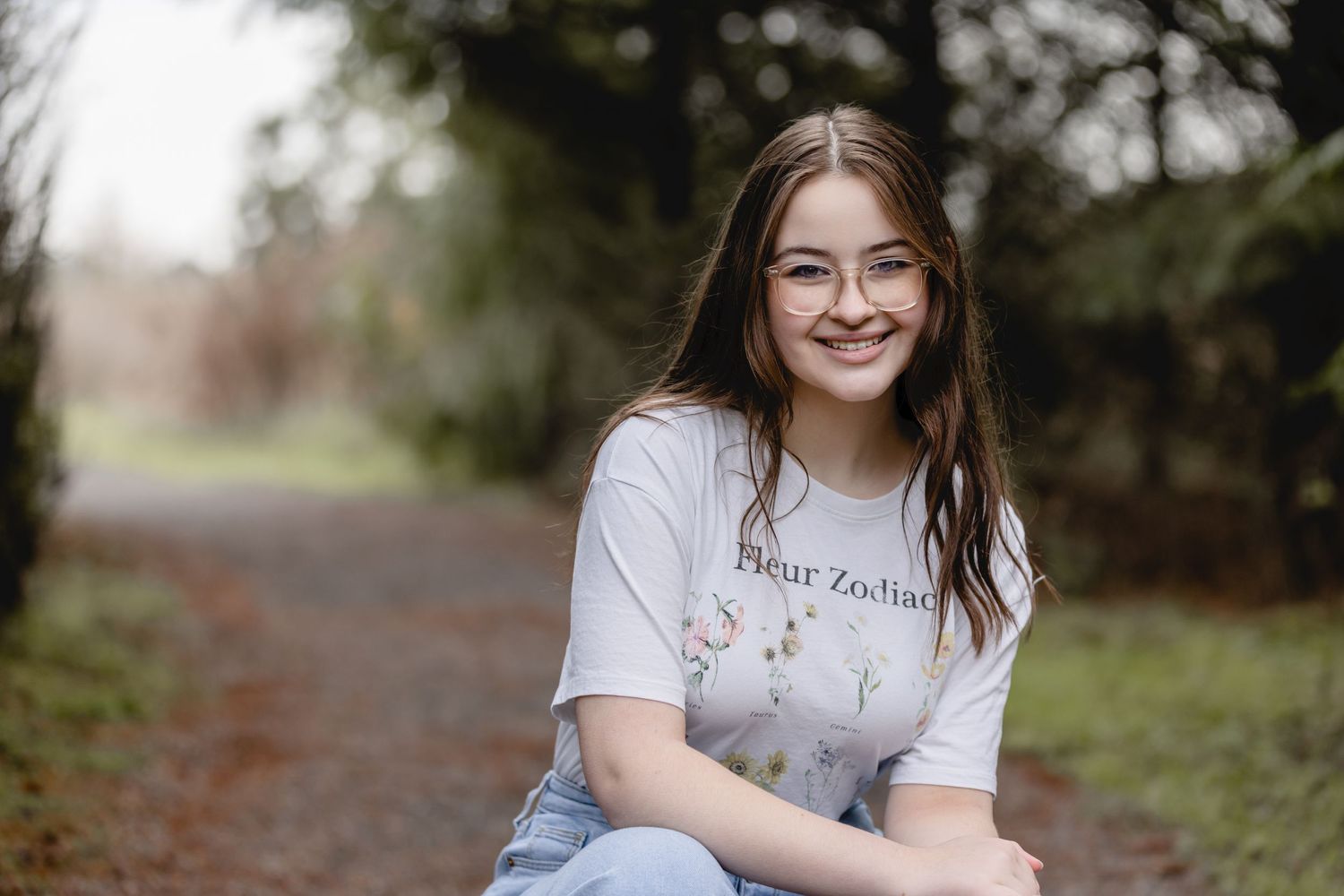 A person wearing a light gray t-shirt and jeans sits on a path surrounded by greenery while smiling at the camera.