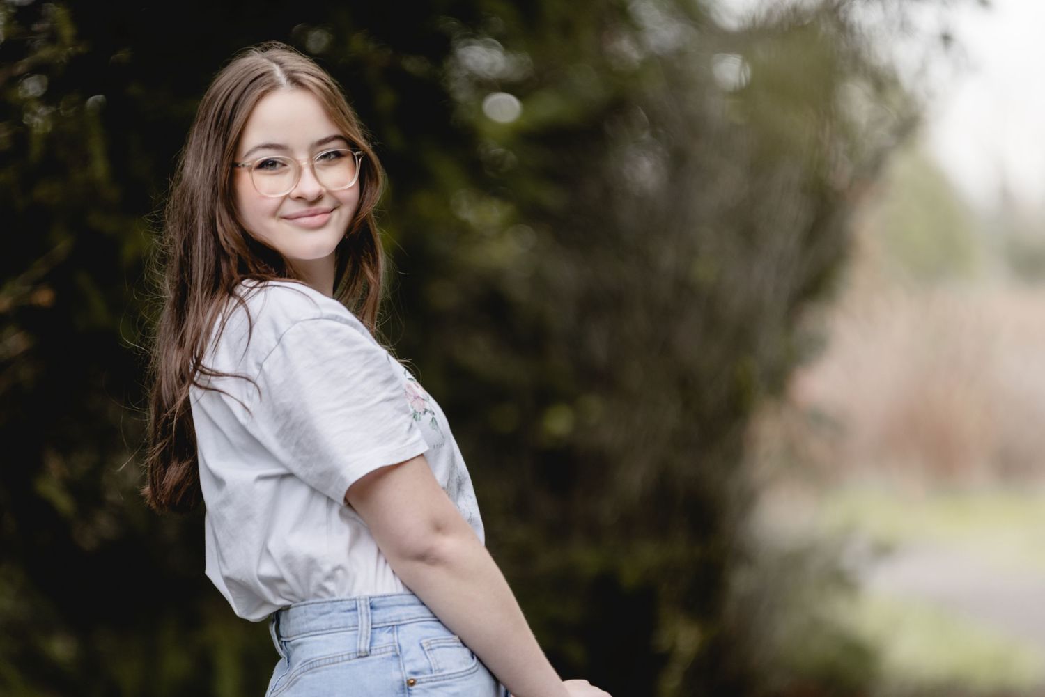 Young woman wearing a white shirt and denim shorts smiles while standing outdoors against a blurred natural background.