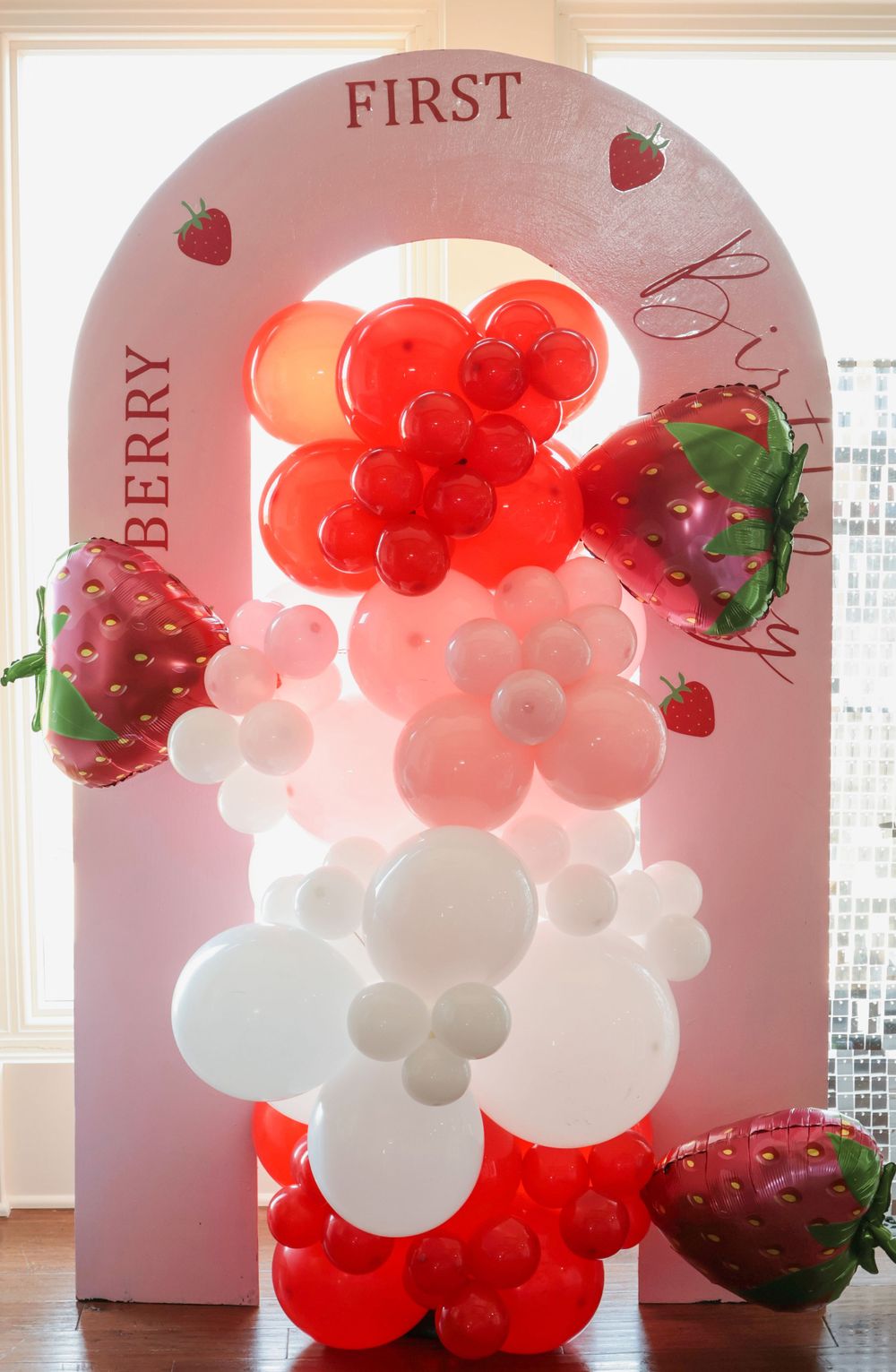 Red and white balloon arch with strawberry decorations against pink wall.