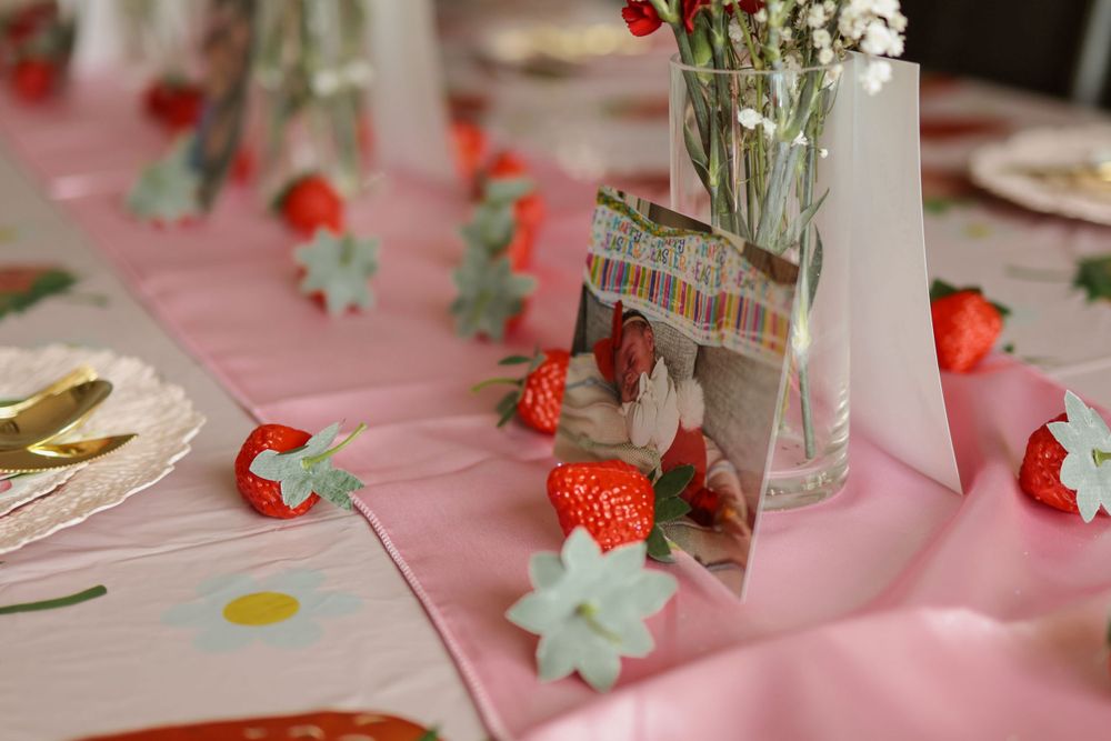 Table setting with scattered flowers and strawberry decorations on pink tablecloth.