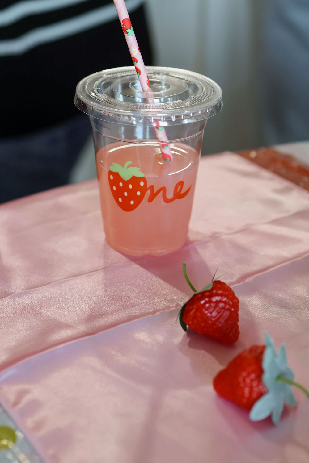 Pink drinks in clear plastic cups with strawberry decorations and straws on a pink tablecloth.