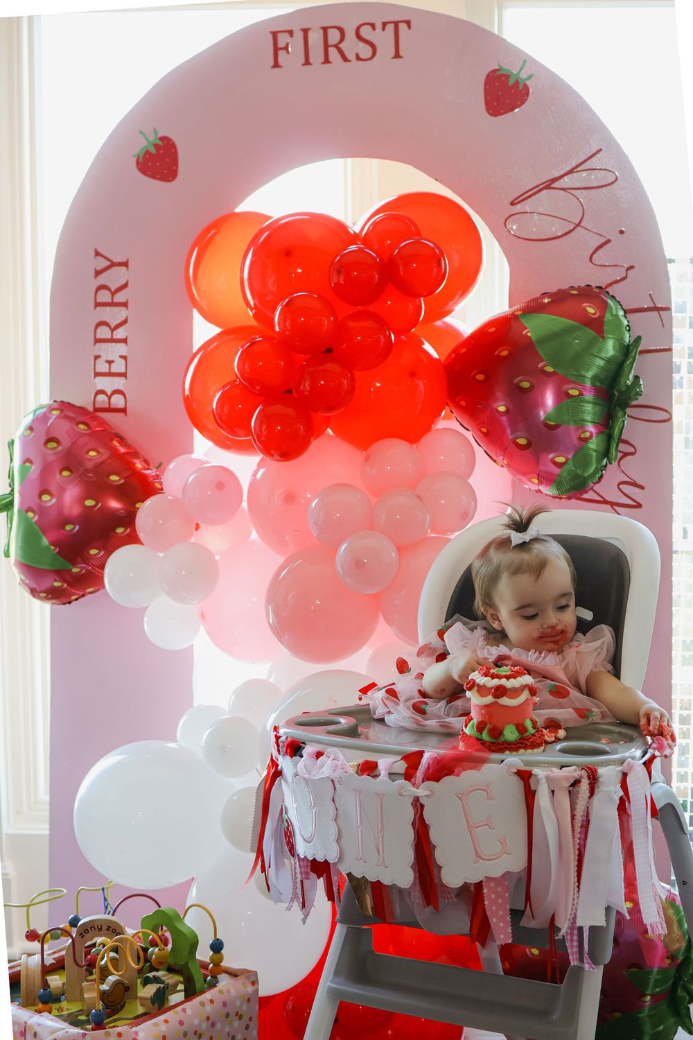 Red and white balloon decorations with strawberry theme create a festive first birthday party atmosphere.
