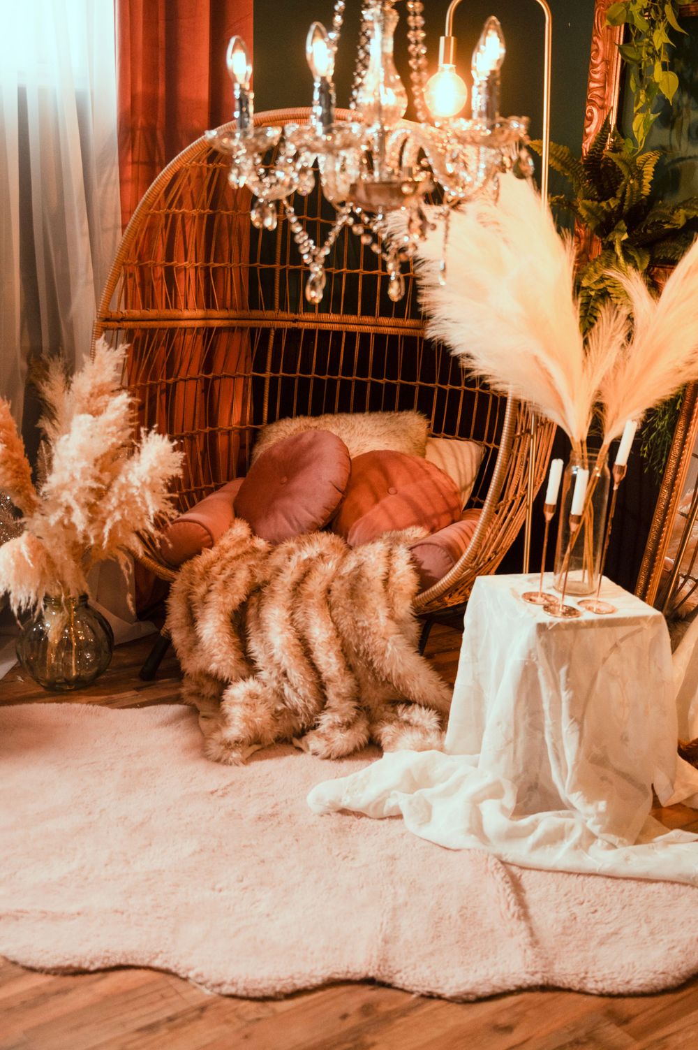Cozy boho room decor with crystal chandelier, wicker chair, white fur rug, and pampas grass in vases.