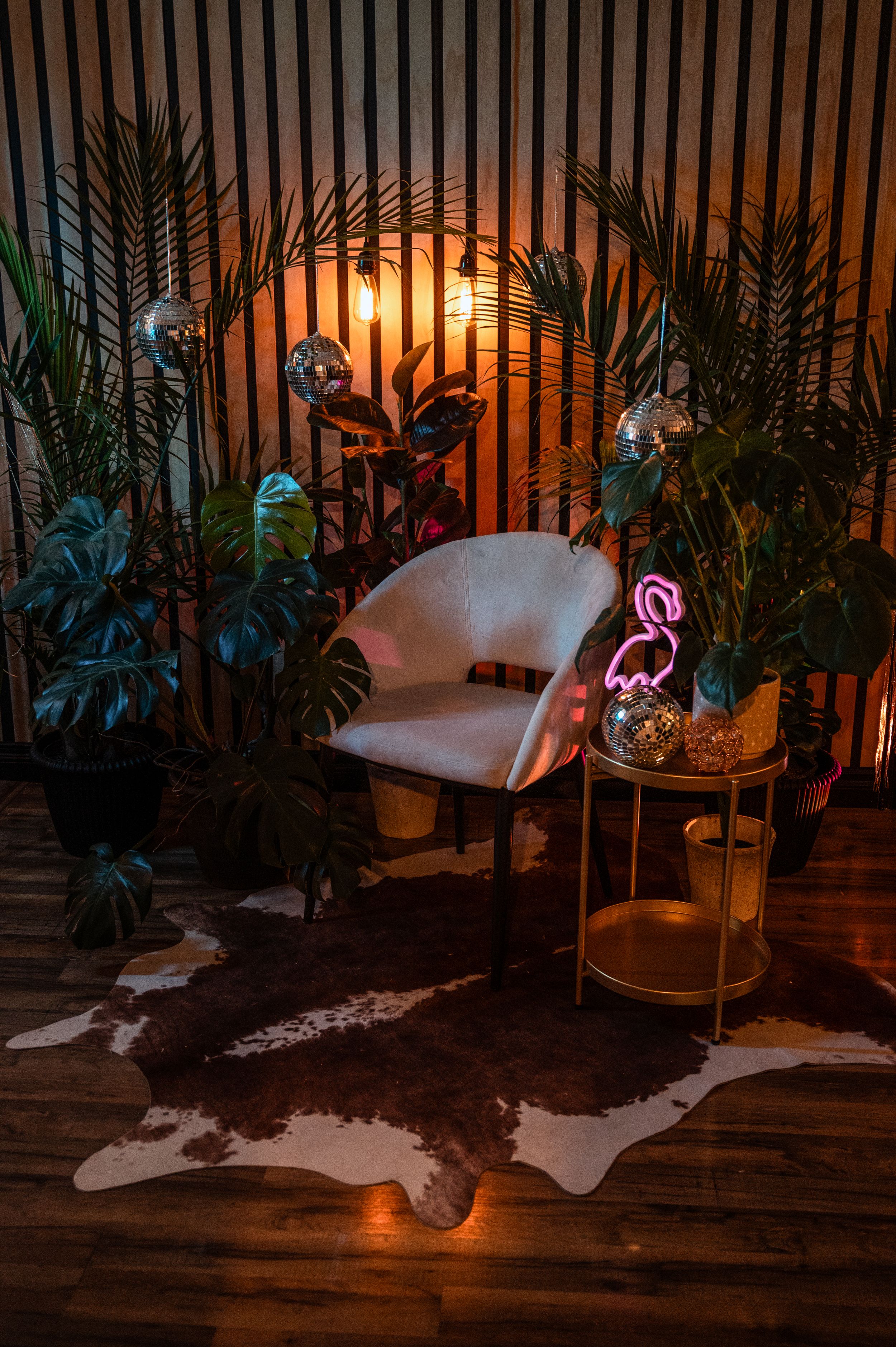 Cozy bohemian interior with white chair, palm plants, neon flamingo light and animal hide rug on wooden floor at sunset.
