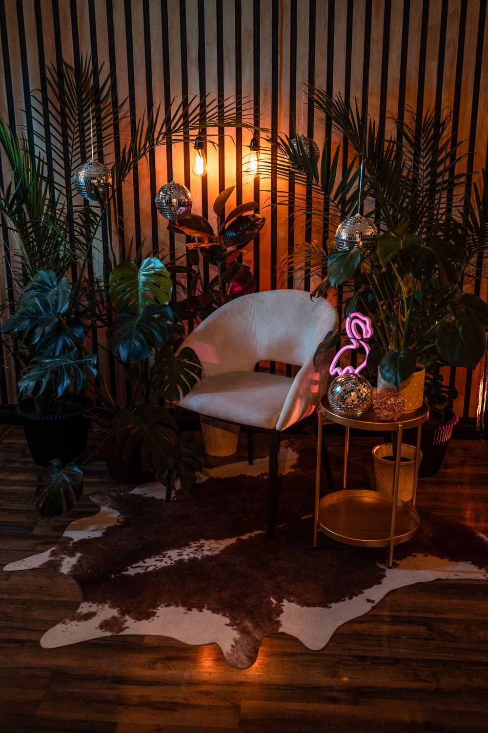 Cozy bohemian interior with white chair, palm plants, neon flamingo light and animal hide rug on wooden floor at sunset.