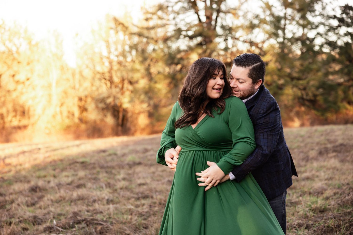 A couple embraces in a sunset-lit field while wearing a green dress and navy jacket on an autumn afternoon.