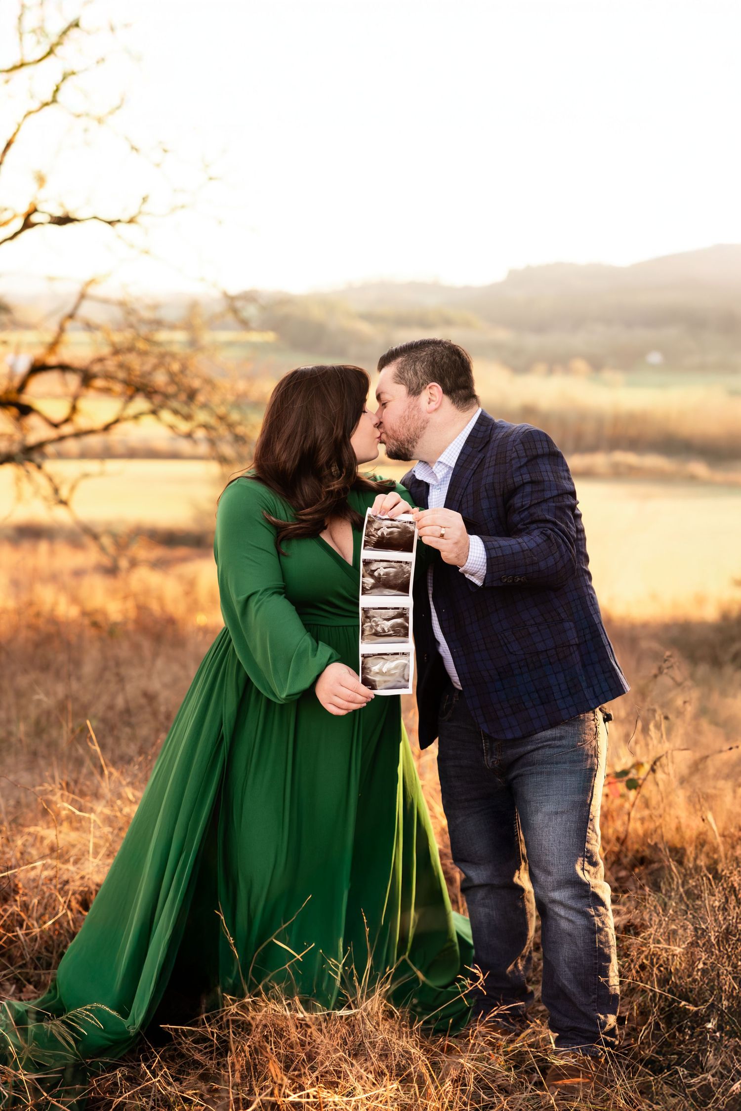 A couple shares a tender moment in a field at sunset while holding ultrasound photos in a beautiful emerald green dress.