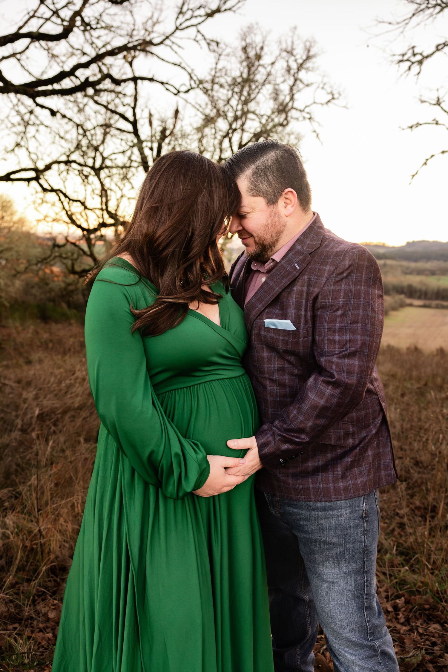 A romantic maternity photo outdoors at sunset featuring an emerald green dress and plaid blazer.