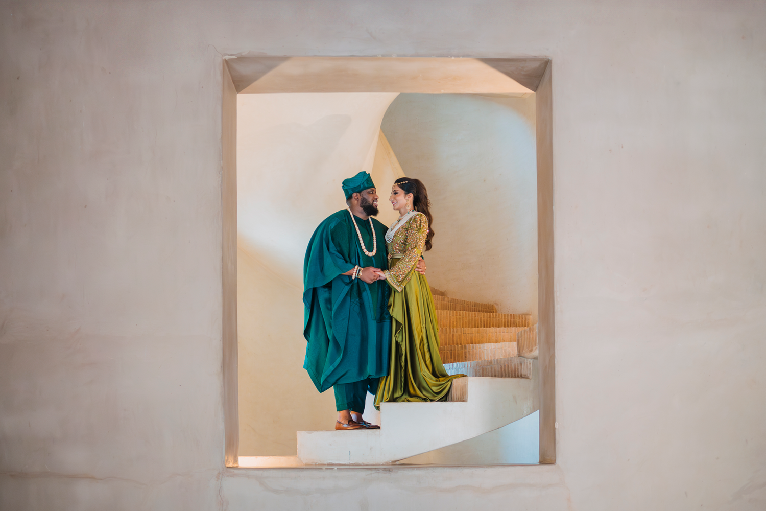 A couple in graduation gowns, one wearing teal and one in yellow, stand together on light-colored stairs.