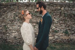 Dans le parc du chateau, les mariés avec leurs lunettes de soleil se tiennent face à face en se regardant costume vert et couronne de fleurs