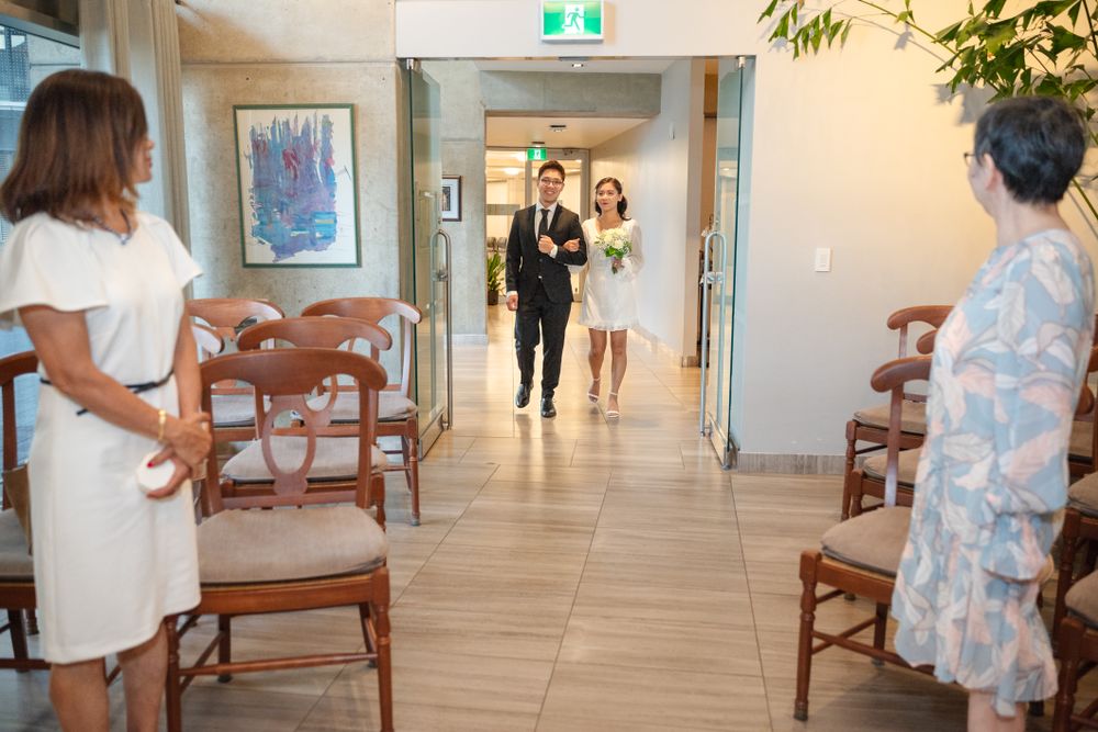 A couple enters a hallway during a wedding ceremony while guests in white dresses sit on wooden chairs.