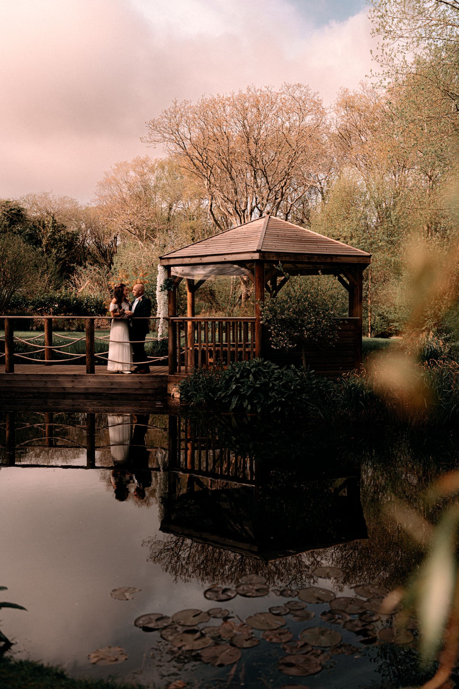 Rustic wooden gazebo with reflection in calm pond water at sunset, surrounded by autumn trees and lily pads.