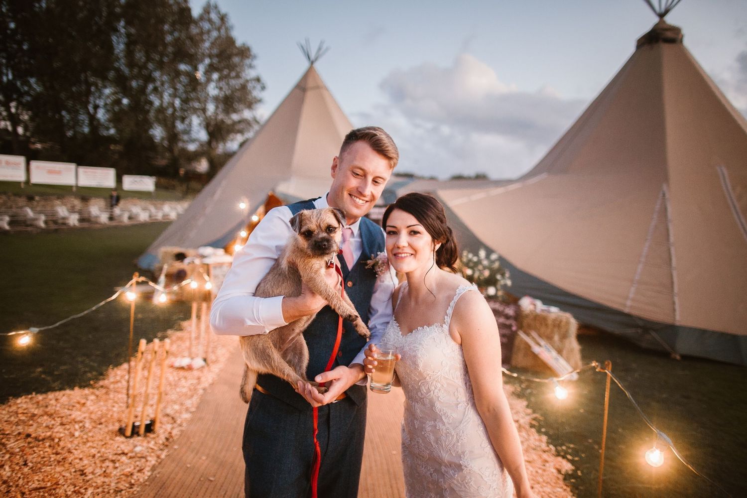 bride and groom stand in front of their wedding tipi holding their beloved border terrier dog