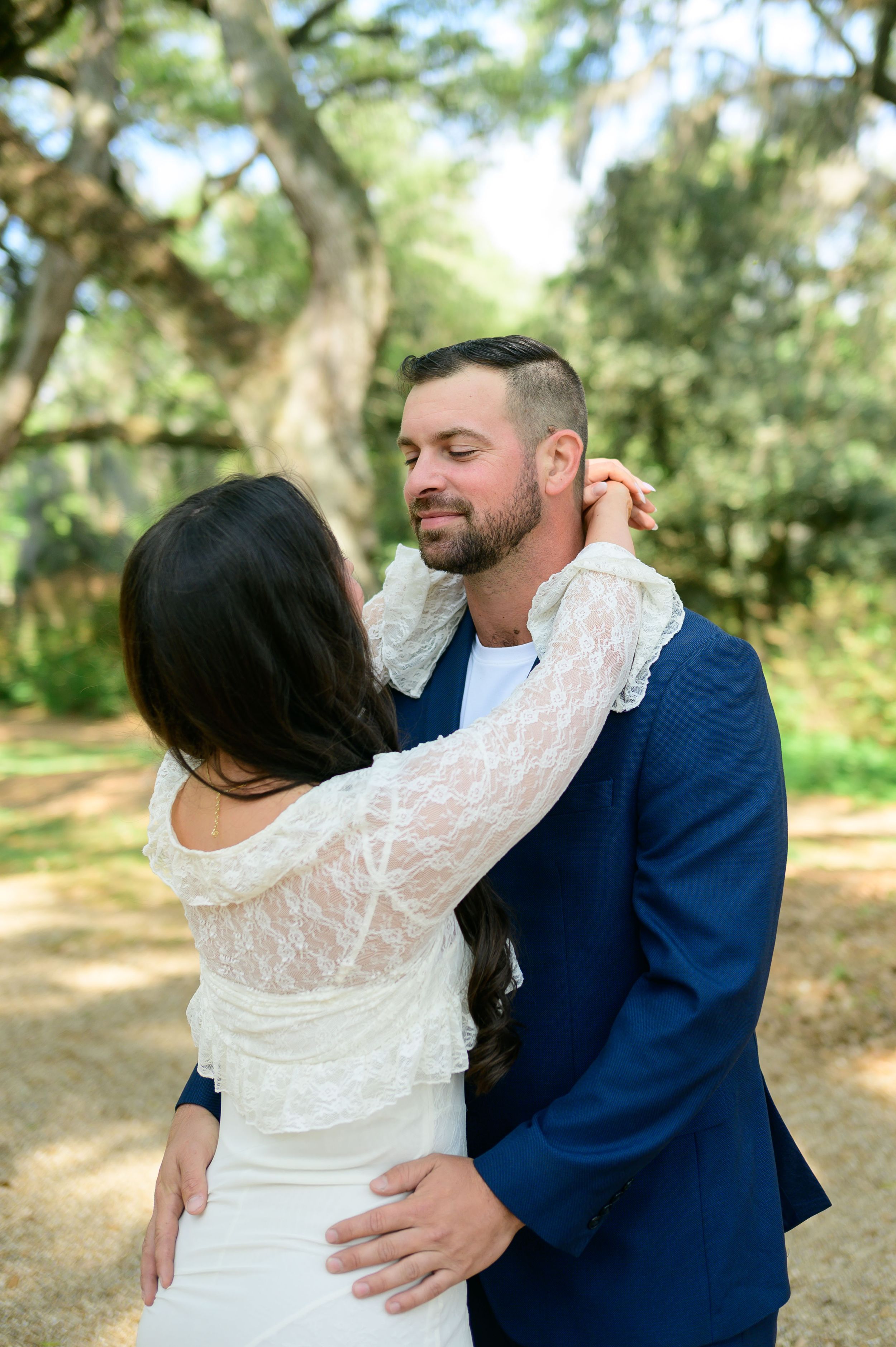 Bride in lace dress embraces groom in navy suit outdoors under oak trees at a romantic wedding ceremony.