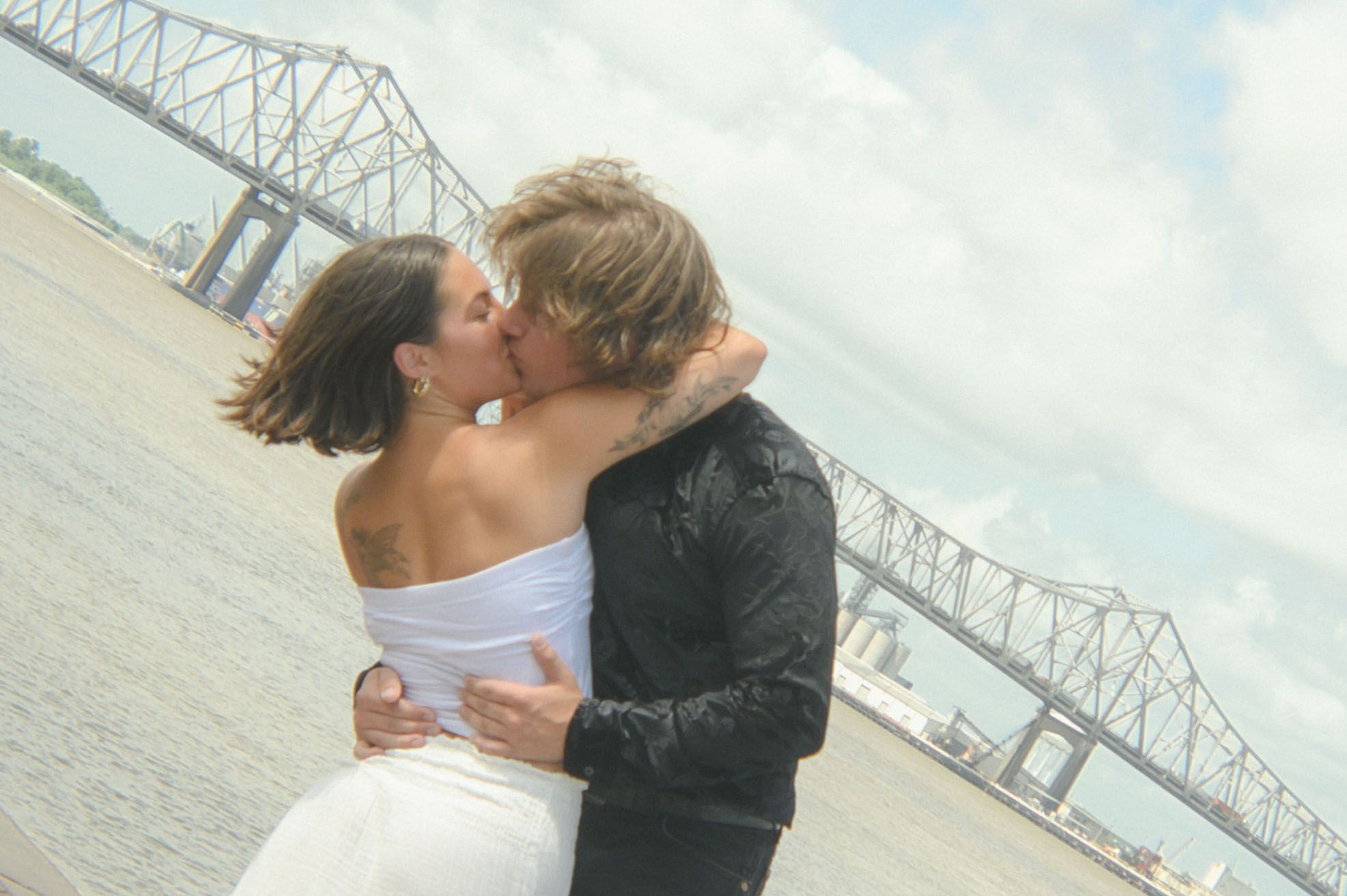 Couple embraces with bridge visible in background during romantic waterfront moment.