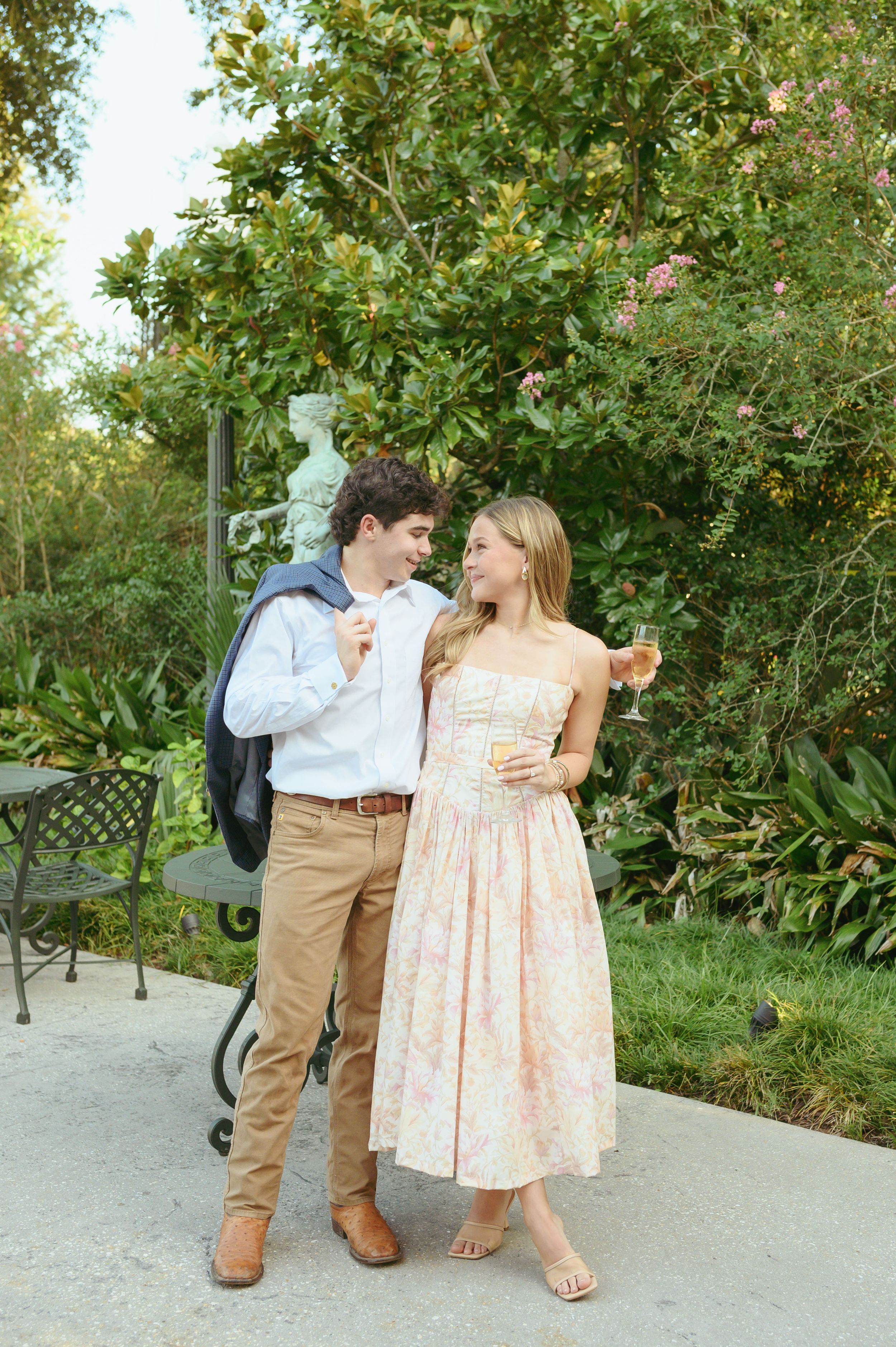 A couple sharing drinks together in a garden setting wearing summer attire with trees and greenery in the background.