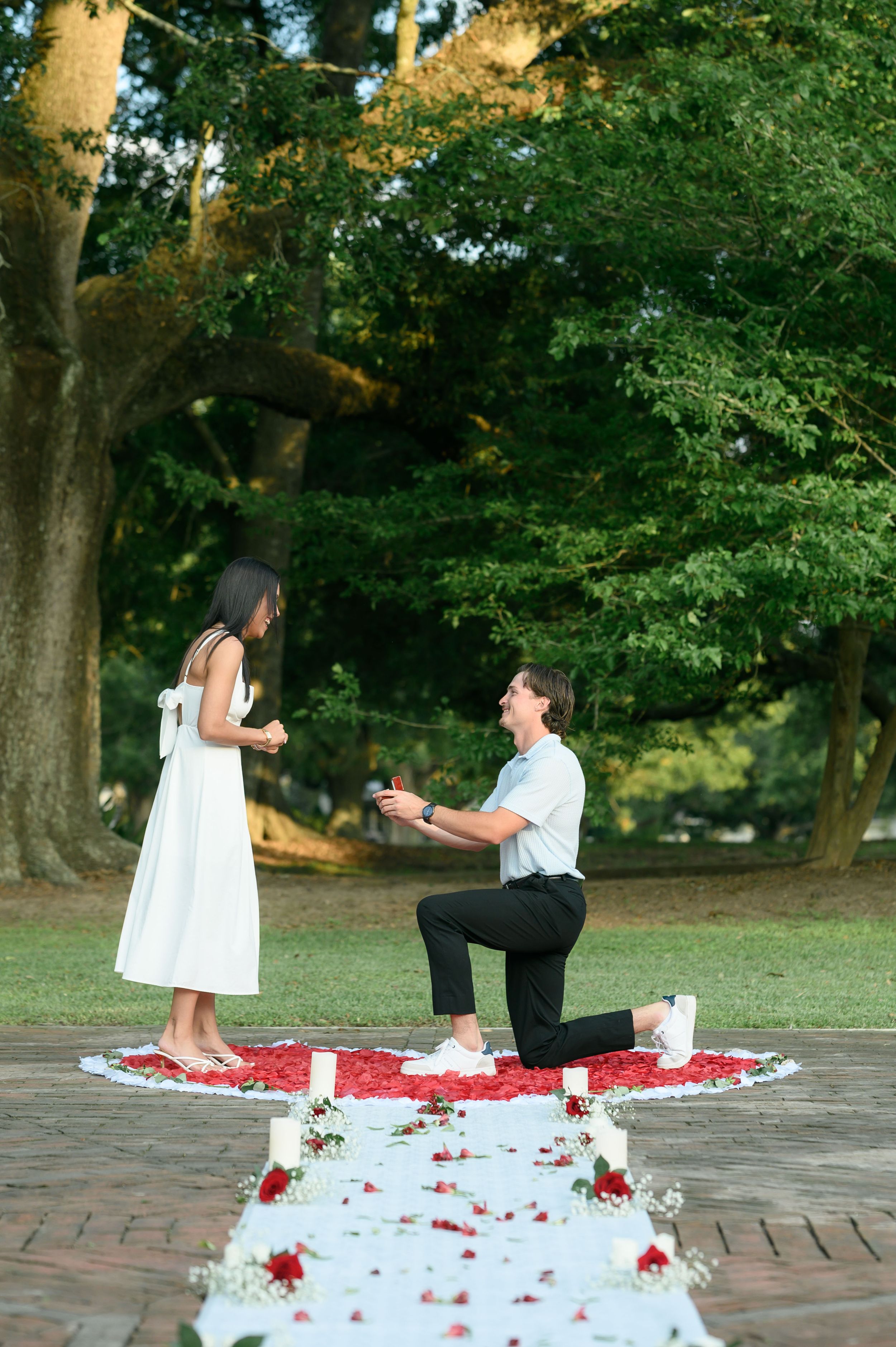 Man kneeling on one knee proposing to woman in white dress on rose petal path outdoors.