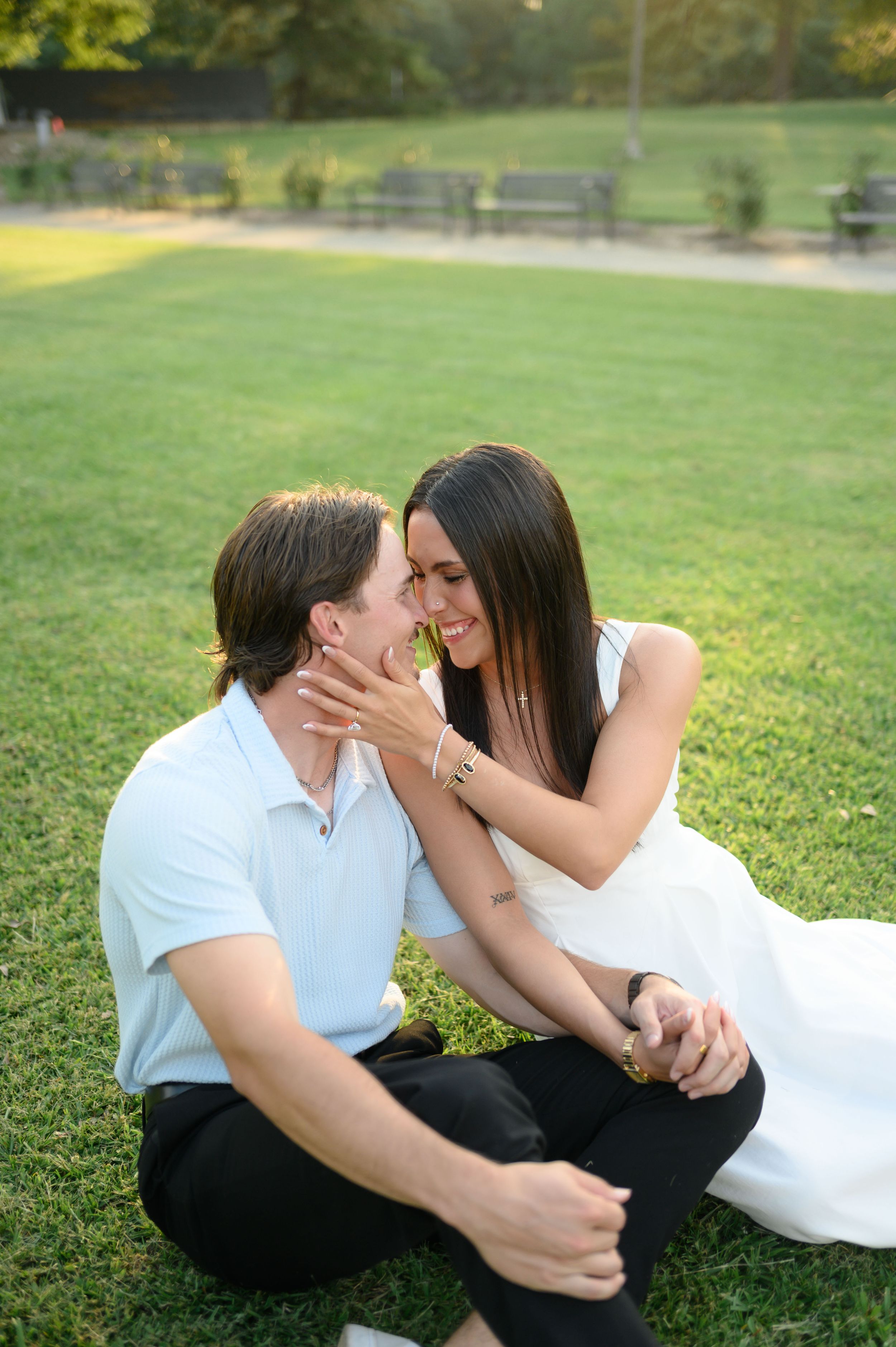 Happy couple sitting on grass, woman cradling mans face, sharing romantic moment outdoors.