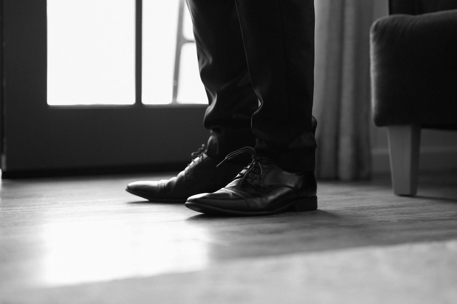 Close-up black and white photo of formal dress shoes on a hardwood floor near a window.