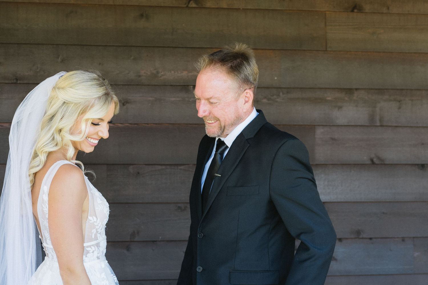 A tender moment between a bride in white dress and someone in a dark suit against a wooden wall.